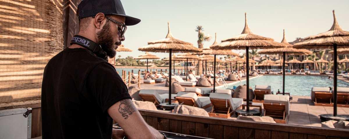 DJ at the decks in the poolside DJ booth at Cook's Club El Gouna hotel in Egypt. View of sunloungers, beanbags and thatched parasols by the pool.