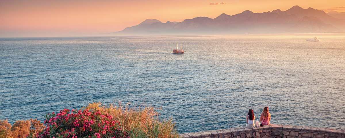 Two women sitting on a sea wall with their backs to the camera looking out across the sea. There are boats on the water and a misty mountainous coastline in the background